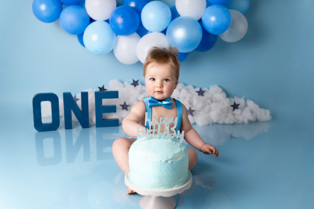 baby boy on his first birthday with a big blue birthday cake and balloons, photographed by Newborn Photographer in Lichfield staffordshire